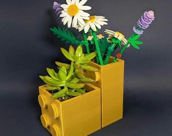 May include: A yellow plastic building block planter with two tiers, containing a green succulent plant and a bouquet of artificial flowers including white daisies with yellow centers and purple lavender-like stems made from interlocking bricks.