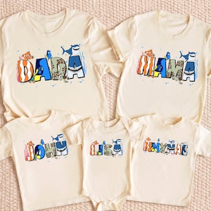 May include: A set of five cream-colored short-sleeve shirts and one bodysuit featuring colorful graphic lettering. The designs include cartoon fish and sea creature motifs integrated into the letters spelling DADA, MAMA, JOHN, LISA, and NNYNAC.