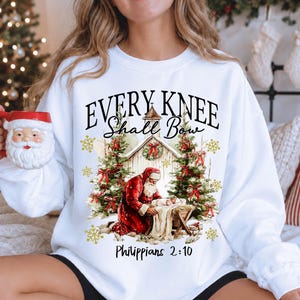 May include: A white long-sleeve sweatshirt featuring a graphic of Santa Claus kneeling before a manger. The text reads "EVERY KNEE Shall Bow" and "Philippians 2:10". The design includes two Christmas trees, gold snowflakes, and a rustic building.