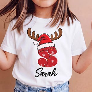 Pode incluir: Uma t-shirt branca de manga curta com uma letra S vermelha grande decorada com flocos de neve brancos e luzes coloridas. A letra é rematada com um gorro de Pai Natal vermelho e branco e hastes de rena castanhas. O nome Sarah está impresso abaixo.