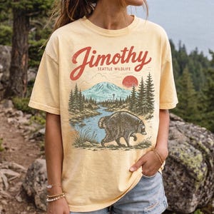 A woman wearing a yellow t-shirt with a graphic design of a bear and a mountain landscape, standing in front of a forested area.
