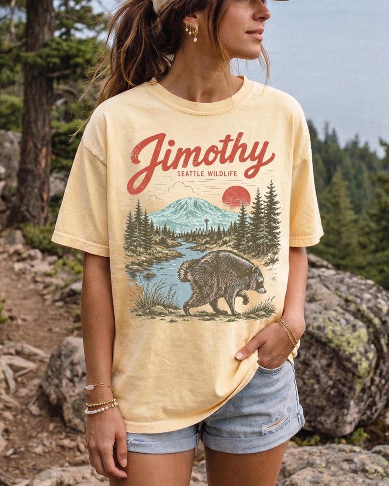 A woman wearing a yellow t-shirt with a graphic design of a bear and a mountain landscape, standing in front of a forested area.