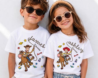 May include: Two white short-sleeved t-shirts featuring Toy Story character graphics. One shirt displays Woody riding Bullseye with the name James, and the other shows Jessie riding Bullseye with the name Ashley, both surrounded by colorful space icons.
