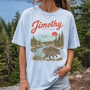 A woman wearing a light blue t-shirt with a graphic design of a bear and a mountain landscape, standing in front of a forested area.