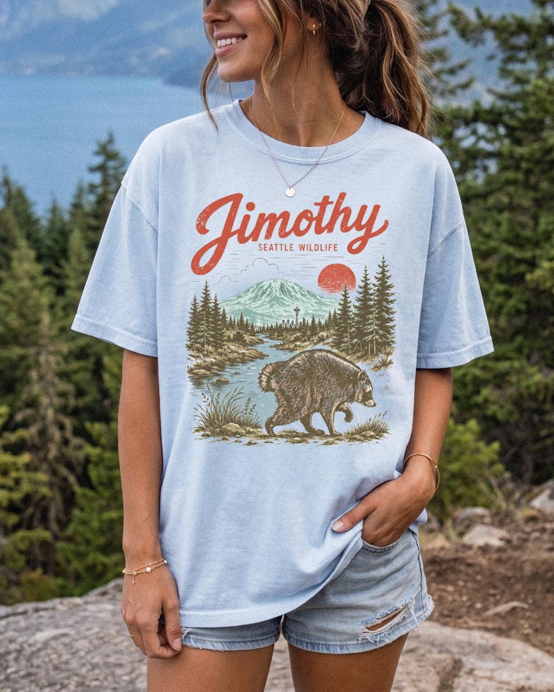 A woman wearing a light blue t-shirt with a graphic design of a bear and a mountain landscape, standing in front of a forested area.