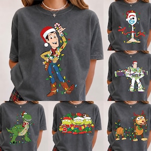 May include: A collection of six charcoal gray t-shirts featuring various Toy Story characters decorated with Christmas lights. The designs include Woody, Forky, Buzz Lightyear, Rex, the Aliens, and Slinky Dog, all wearing festive holiday accessories.