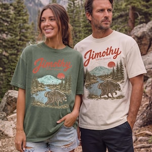 A woman and a man standing together in a forest, both wearing t-shirts with a graphic design featuring a bear and mountains.