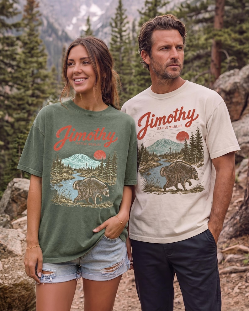 A woman and a man standing together in a forest, both wearing t-shirts with a graphic design featuring a bear and mountains.