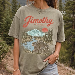 A woman wearing a t-shirt with a graphic design of a mountain, river, and bear, standing in a forested area.