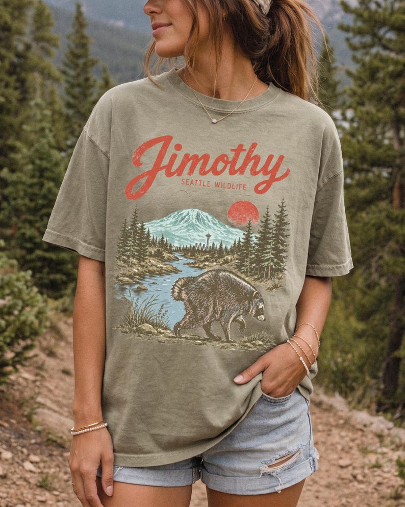 A woman wearing a t-shirt with a graphic design of a mountain, river, and bear, standing in a forested area.