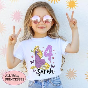 May include: A white short-sleeved t-shirt featuring a graphic of the character Rapunzel with long blonde hair, a purple dress, and a green chameleon. The design includes a large purple number 4, purple butterflies, and the name Sarah in black.