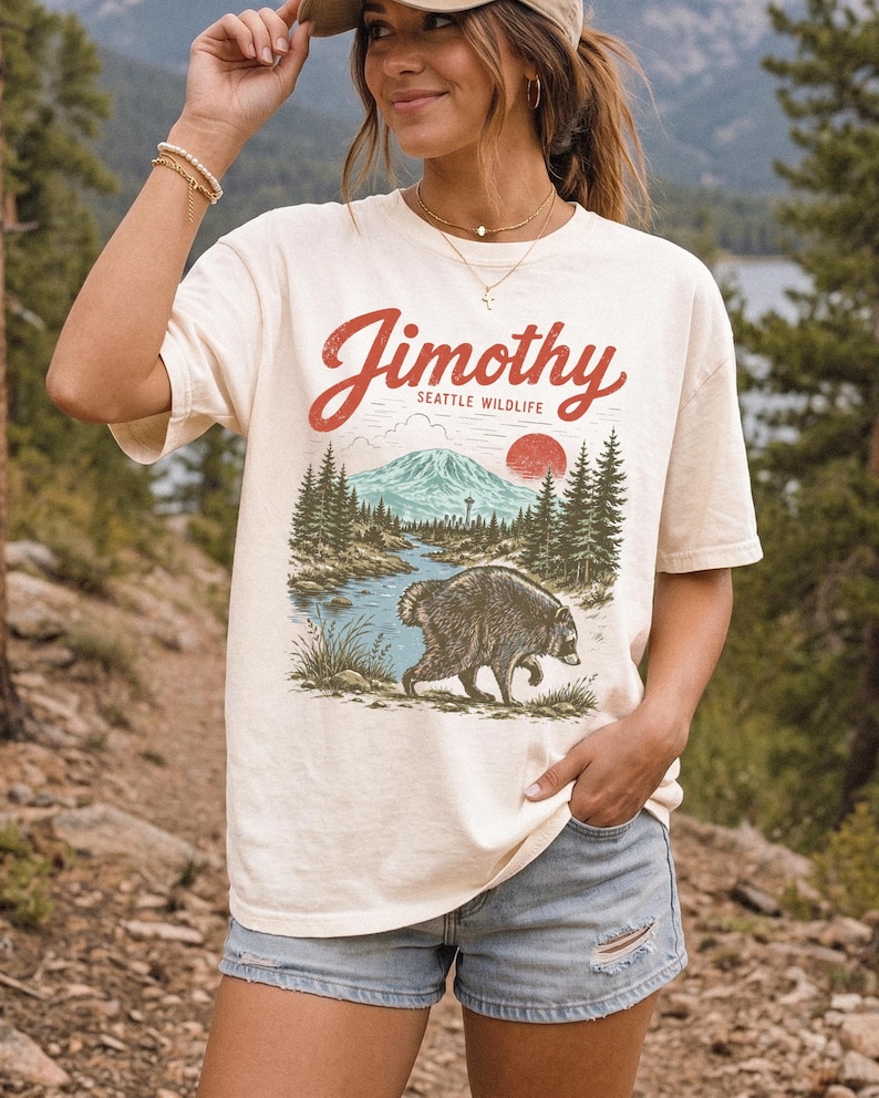 A woman wearing a white t-shirt with a graphic design of a bear and mountains, standing in a forested area.