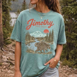 A woman wearing a green t-shirt with a graphic of a bear and a mountain landscape, standing in a forested area.