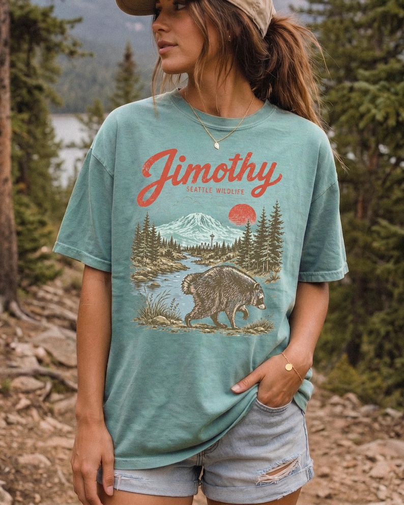 A woman wearing a green t-shirt with a graphic of a bear and a mountain landscape, standing in a forested area.
