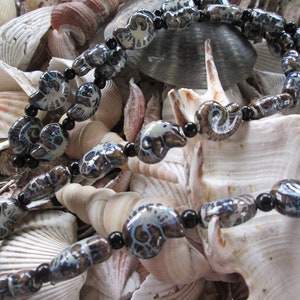 May include: A necklace made of black and blue ceramic beads with a swirl pattern. The beads are shaped like ammonites and are strung on a black cord. The necklace is displayed on a bed of seashells.