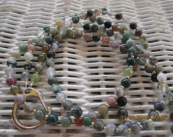 May include: A multi-strand necklace made of green, brown, and white agate beads with a silver clasp. The necklace is photographed on a white wicker surface.