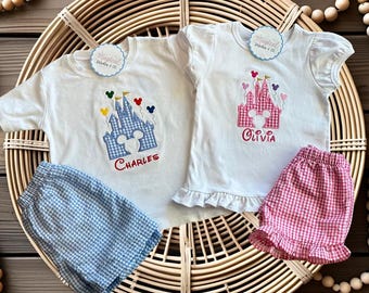 May include: Two white t-shirts with castle embroidery and matching shorts. One shirt has a blue castle with "Charles" embroidered below, the other has a pink castle with "Olivia". The shorts are blue and pink gingham.