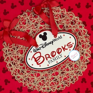 Op de afbeelding: Een decoratief familieornament met de tekst "Brooks Family" in het rood, met een Mickey Mouse silhouet en de tekst "Walt Disney World". Het ornament staat op een geweven, cirkelvormige basis met rode linten, tegen een rode achtergrond met Mickey Mouse iconen.