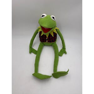 May include: A green plush frog doll with long, thin limbs and a pointed collar. The frog wears a red and navy blue plaid vest with two small gold buttons on the front. It has large white eyes with black pupils and a wide, open red mouth.