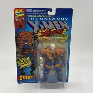 May include: An X-Men X-Force Grizzly action figure in its original packaging. The figure is orange and blue, holding a silver weapon. The packaging features the Marvel Comics logo and the text "CRUSHING POWER HAMMERS!" The figure is in a clear plastic case.