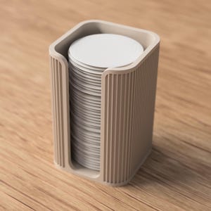 May include: A beige, rectangular coaster holder with a stack of white coasters. The holder has a ribbed design on the sides and a curved top. The coasters are round and neatly stacked inside the holder.