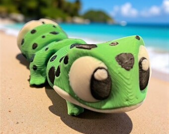 May include: A green and black cartoon-style lizard figurine with large, round eyes and a segmented body. The lizard is lying on a sandy beach with a blurred ocean and sky background. The figurine has a textured surface.