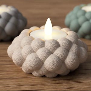 May include: Three decorative candle holders with a textured, bubble-like design. The front candle holder is off-white, with two others in the background, one gray and one teal. Each holds a lit, battery-operated candle, creating a warm glow.