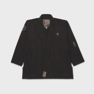May include: A black martial arts gi with the brand name "WAZAF" printed on the sleeve. The uniform features a collar and is made of a robust material. Patches with the brand logo are visible on the chest and shoulder.