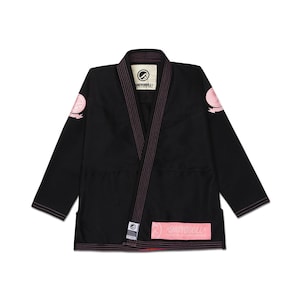 May include: A black martial arts gi jacket featuring pink contrast stitching and trim. The garment includes circular pink patches on each shoulder, a rectangular pink patch with the text SHOYOROLL on the lower front, and a branded inner neck label.