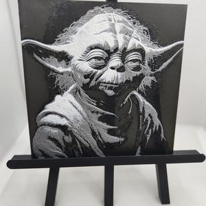 Pode incluir: Retrato a preto e branco de Yoda de Star Wars numa pequena tela. A obra de arte é exibida num cavalete preto em miniatura. A imagem apresenta sombreamento e textura detalhados.