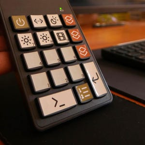 Könnte beinhalten: Eine schwarze, kompakte Tastatur mit weißen Tasten und verschiedenen Symbolen, darunter Strom-, Helligkeits- und Mediensteuerungen. Die Tastatur wird vor einem Computermonitor mit einer grünen Landschaft gehalten.