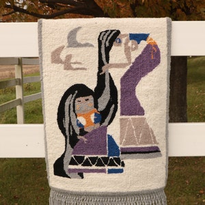 May include: A woven wall hanging with a fringe border features a colorful design of two figures in purple, blue, and black against a white background.