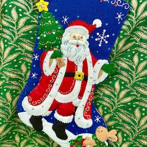 Könnte beinhalten: Ein blauer Filz-Weihnachtsstrumpf mit einem Weihnachtsmann- und Weihnachtsbaummotiv. Der Strumpf ist mit Pailletten und Perlen verziert. Der Weihnachtsmann hält einen Weihnachtsbaum und trägt einen roten Anzug mit weißem Bart und weißem Pelzbesatz. Ein kleines braunes Kaninchen sitzt am unteren Ende des Strumpfes.