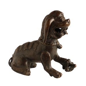 May include: A bronze sculpture of a stylized lion with a detailed mane and a curled tail. The lion is sitting with its head turned to the side and its mouth slightly open.