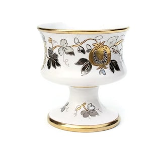 Puede incluir: Un gobelet de cerámica blanca con un borde dorado y un diseño floral en oro y plata. El gobelet tiene una base de pedestal.