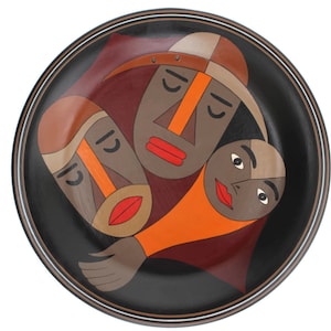 May include: A round black plate with a white rim features three stylized faces in orange, brown, and red. The faces are painted in a geometric style and have closed eyes.