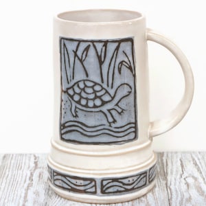 Puede incluir: Una jarra de cerveza de cerámica blanca con un diseño de tortuga marrón y olas de agua azules. La jarra tiene un asa y una base con un patrón decorativo.