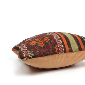 May include: A colorful patterned pillow with a tan suede backing. The pillow is made of woven fabric with stripes and geometric designs in shades of red, orange, green, brown, and black.