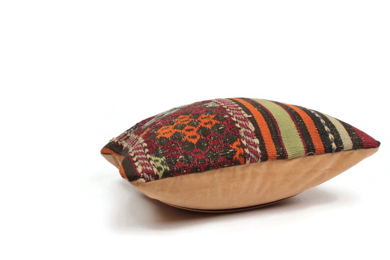 May include: A colorful patterned pillow with a tan suede backing. The pillow is made of woven fabric with stripes and geometric designs in shades of red, orange, green, brown, and black.