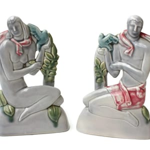 May include: A pair of ceramic figurines depicting a man and a woman, each holding a dove. The figurines are painted in shades of blue, green, and pink, and they are sitting on a base that resembles a rock.