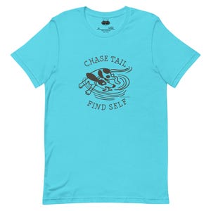 May include: A turquoise t-shirt featuring a brown graphic of two dogs playing in water. The text "CHASE TAIL" is arched above the dogs, and "FIND SELF" is below. The shirt has a crew neck and short sleeves.