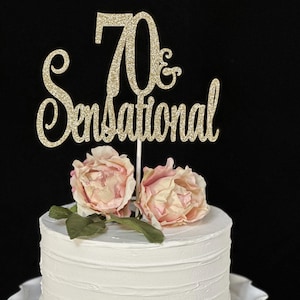 May include: Gold glitter cake topper with the text "70 & Sensational" on a white cake with pink flowers.