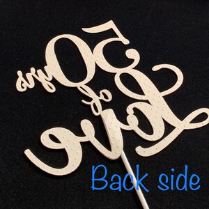 May include: White cake topper with the text "50 and fabulous" in a cursive font. The topper is on a stick and has a dotted pattern.