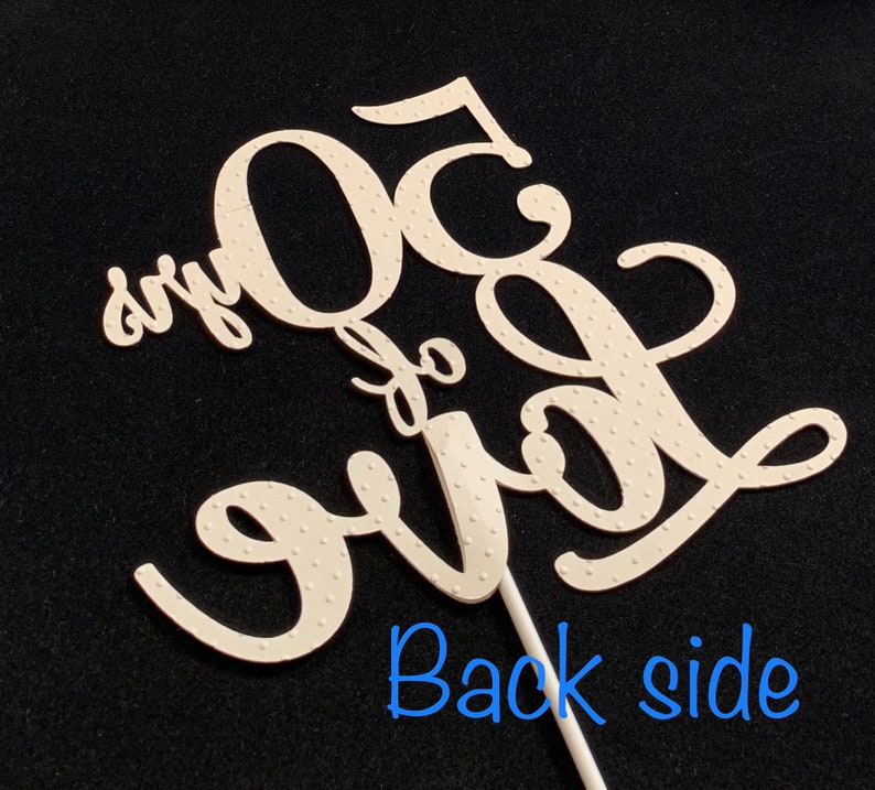 May include: White cake topper with the text "50 and fabulous" in a cursive font. The topper is on a stick and has a dotted pattern.