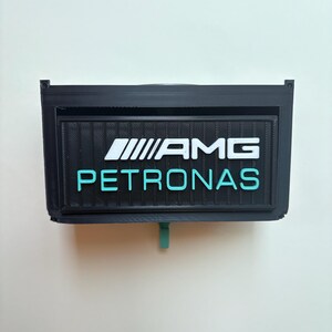 May include: A black rectangular object with the text "AMG PETRONAS" in white and teal. The object has a textured surface and a teal-colored tab extending from the bottom. The AMG logo is prominently displayed.