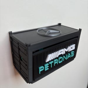 Könnte beinhalten: Eine schwarze, containerförmige Box mit dem AMG Petronas-Logo in Weiß und Türkis. Die Oberseite zeigt ein silbernes Mercedes-Benz-Emblem. Die Box ist an einer weißen Wand befestigt.