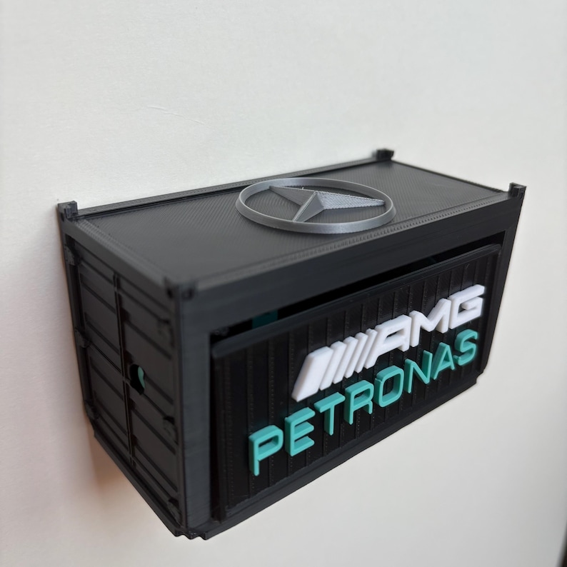 May include: A black container-shaped box with the AMG Petronas logo in white and teal. The top features a silver Mercedes-Benz emblem. The box is mounted on a white wall.