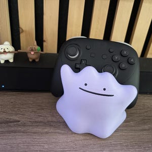 May include: A light purple Ditto figure rests in front of a black game controller. Two small dog figurines with sticks are on a black box. The background features wooden slats.