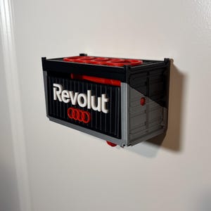 May include: A 3D-printed container-shaped wall mount, featuring the "Revolut" logo in white and the Audi logo in red. The container is primarily black and gray, with red accents. It is mounted on a white wall.