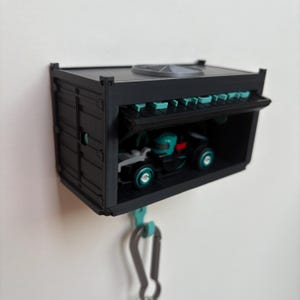 May include: A black miniature shipping container with a hinged door, housing a small toy race car. The container has teal accents and a silver carabiner clip attached. The toy car is black and teal.